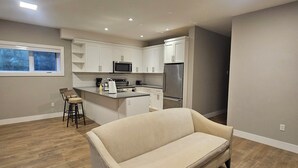 A modern kitchen with white cabinets, a microwave, and a refrigerator. There is a beige sofa and a small table with a colorful tablecloth in the living area.