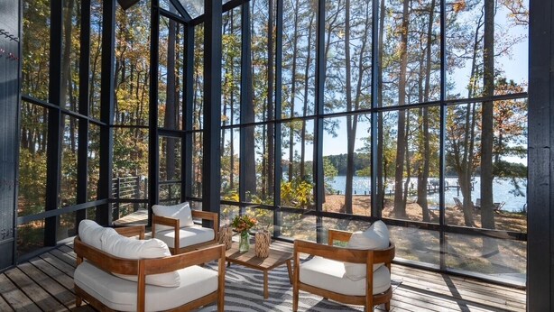 A wooden deck with a sofa and chairs, large windows, and a view of trees and water.