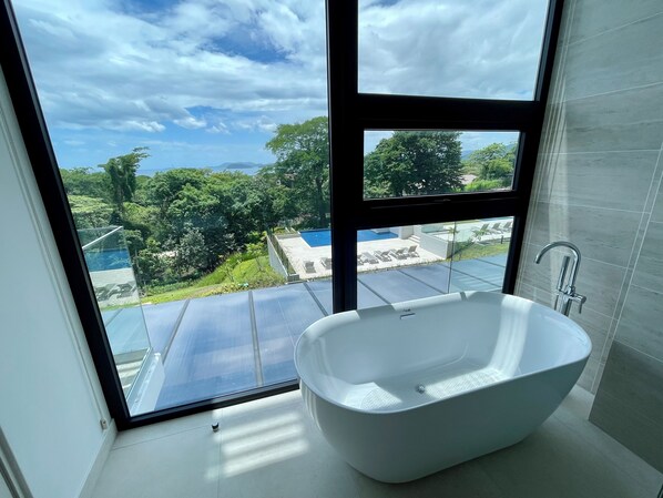 A modern bathroom with a freestanding bathtub, large windows, and a view of a swimming pool and greenery.