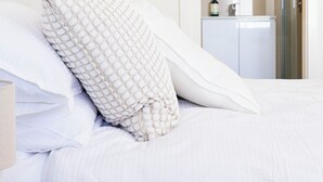 A neatly made bed with white bedding and a textured pillow, a wooden nightstand, and a doorway leading to a bathroom with a mirror and a sink. - Lake Illawarra Family Escape (Lake Illawarra)