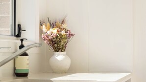 A bathroom with a white sink, a vase with flowers, a clock, and a bottle of hand soap. - Lake Illawarra Family Escape (Lake Illawarra)