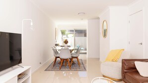 A modern living room with a flat-screen TV, a brown leather sofa, a dining area with a table and chairs, and a large window providing natural light. - Lake Illawarra Family Escape (Lake Illawarra)