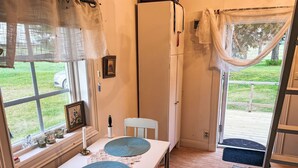 A cozy room with a table set for two, a white chair, a window with curtains, and a view of a grassy area outside.