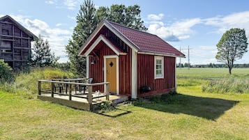 A small red wooden cabin with a porch and a single window, surrounded by a grassy field and trees.