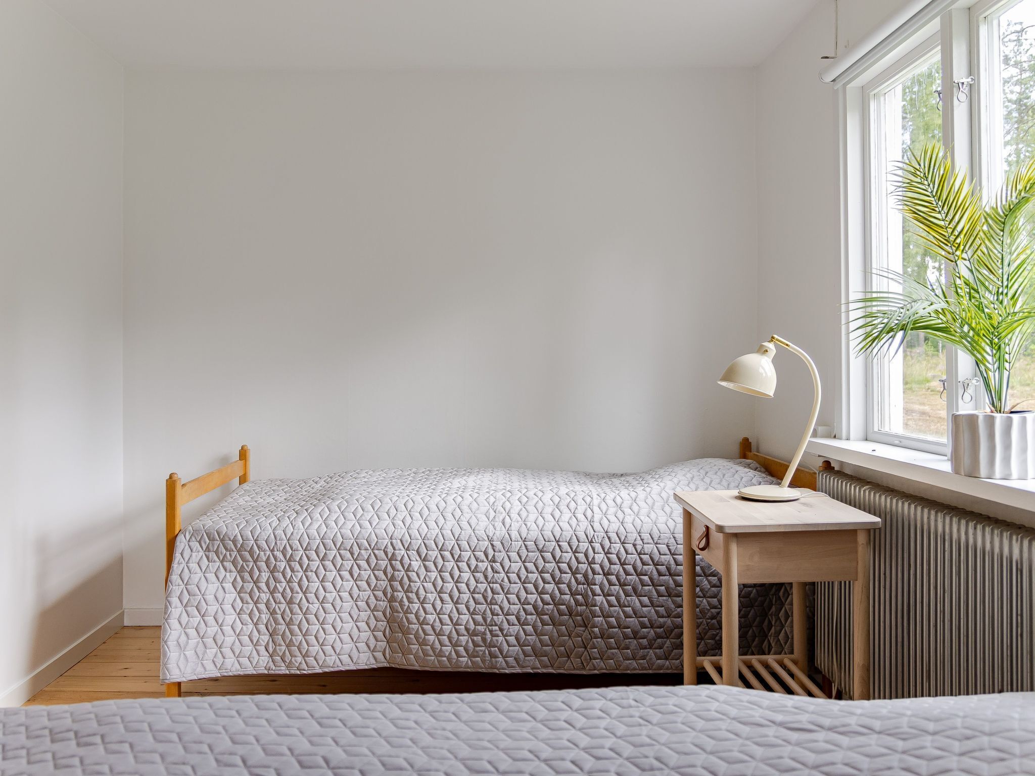 Ett sovrum med en säng, ett litet sängbord, en lampa, ett element och ett fönster med en växt.