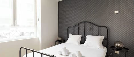 Ein ordentlich bezogenes Bett mit weißen Leinen und Handtüchern, ein schwarzes Metall-Kopfteil, ein kleiner Nachttisch mit Lampe und eine Wand mit dezentem Muster.