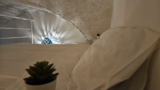 Une chambre avec un lit blanc, une plante en pot et un mur en pierre.