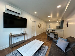 A modern living room with a flat-screen TV, a marble coffee table, a leather sofa, and a dining area with a kitchen in the background.
