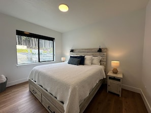 Une chambre avec un grand lit, une table de chevet avec une lampe, une fenêtre donnant sur un paysage verdoyant et un plancher en bois.