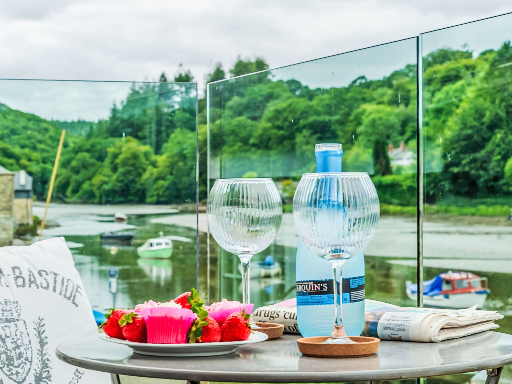 A table with a bottle of water, two glasses, a plate of strawberries, and a newspaper.