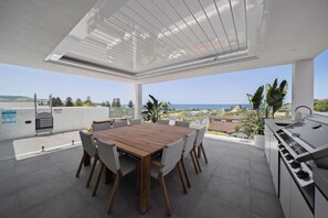Un espace repas extérieur moderne, avec une table et des chaises en bois, un barbecue intégré et une vue sur la mer et le paysage.
