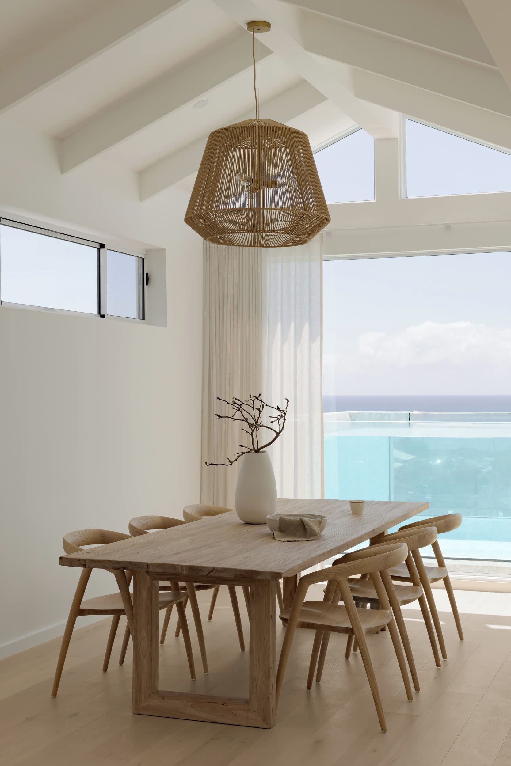 Ein moderner Essbereich mit einem Holztisch und Stühlen, einer Pendelleuchte und Blick auf das Meer durch große Fenster.