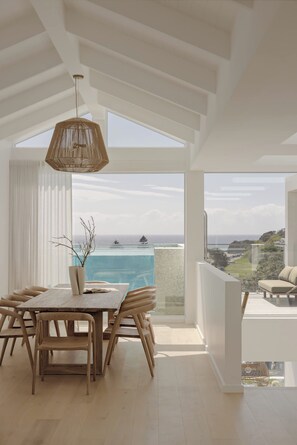 A modern dining area with a view of the ocean, a hanging light fixture, and a wooden table with chairs.