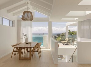 A modern dining area with a wooden table and chairs, a hanging light fixture, and a view of the ocean through large windows.