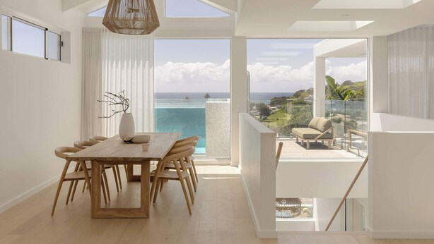 A modern dining area with a wooden table and chairs, a hanging light fixture, and a view of the ocean through large windows.