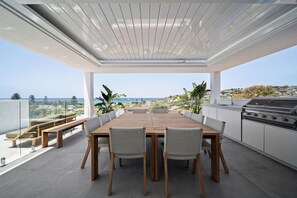 Un espace repas extérieur moderne, doté d’une grande table et de chaises en bois, d’un barbecue et offrant une vue sur l’océan.