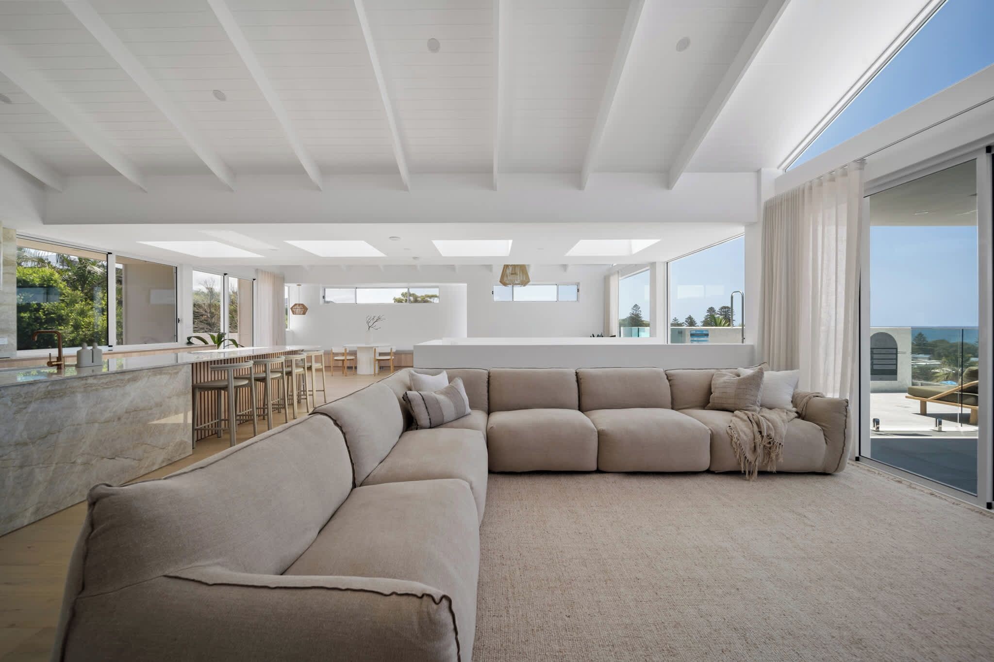 A spacious living area with a large beige sectional sofa, a dining area with a bar, and large windows offering a view of the outdoors.