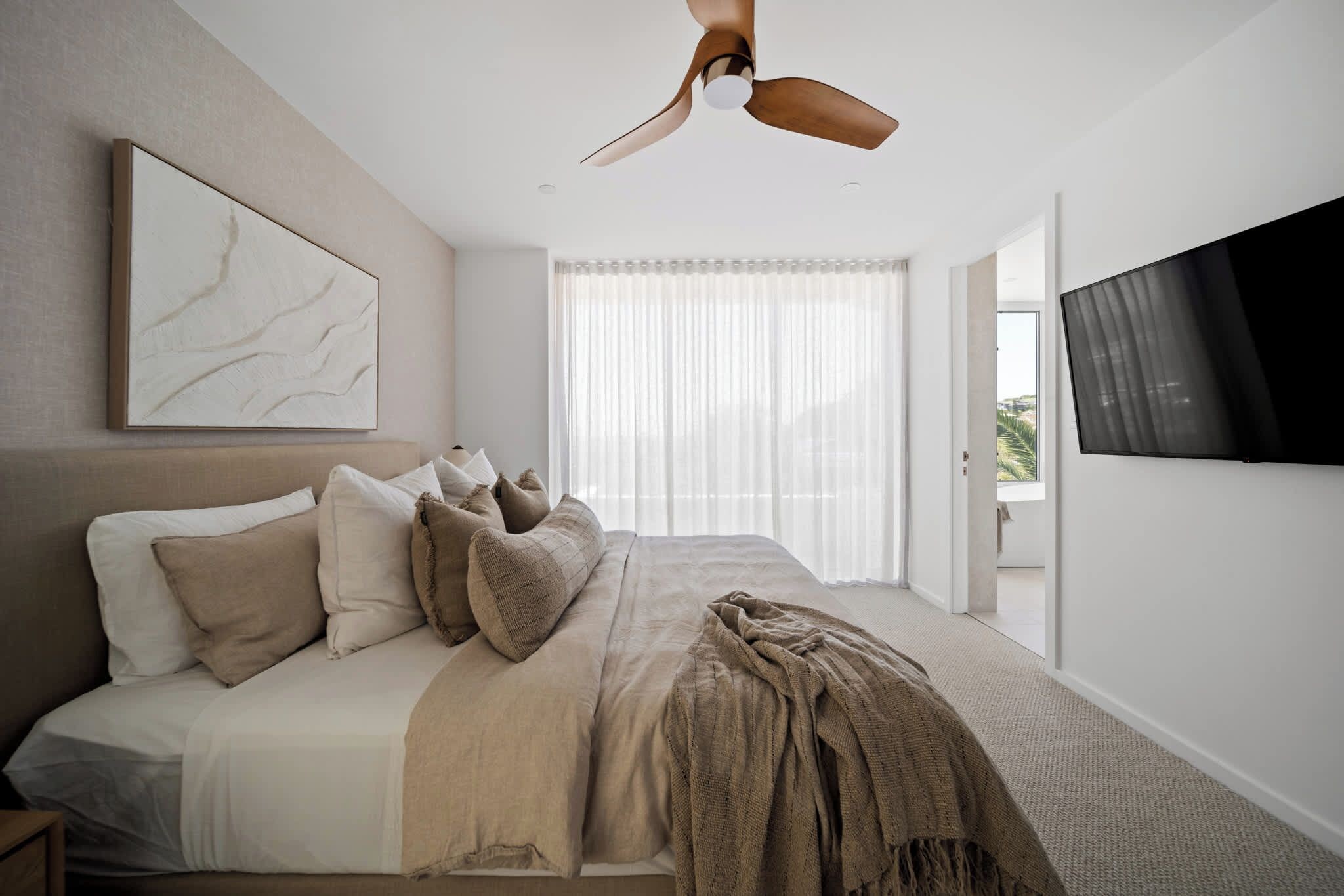 A bedroom with a bed, a ceiling fan, a large window with sheer curtains, and a mounted TV.