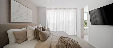 Ein Schlafzimmer mit einem Bett, einem Deckenventilator, einem großen Fenster mit durchsichtigen Vorhängen und einem an der Wand befestigten Fernseher.