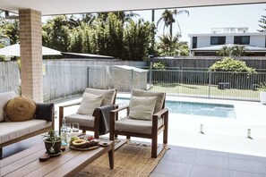 Un patio con dos sillas de madera, una mesa de madera con un mantel tejido, y una piscina al fondo.