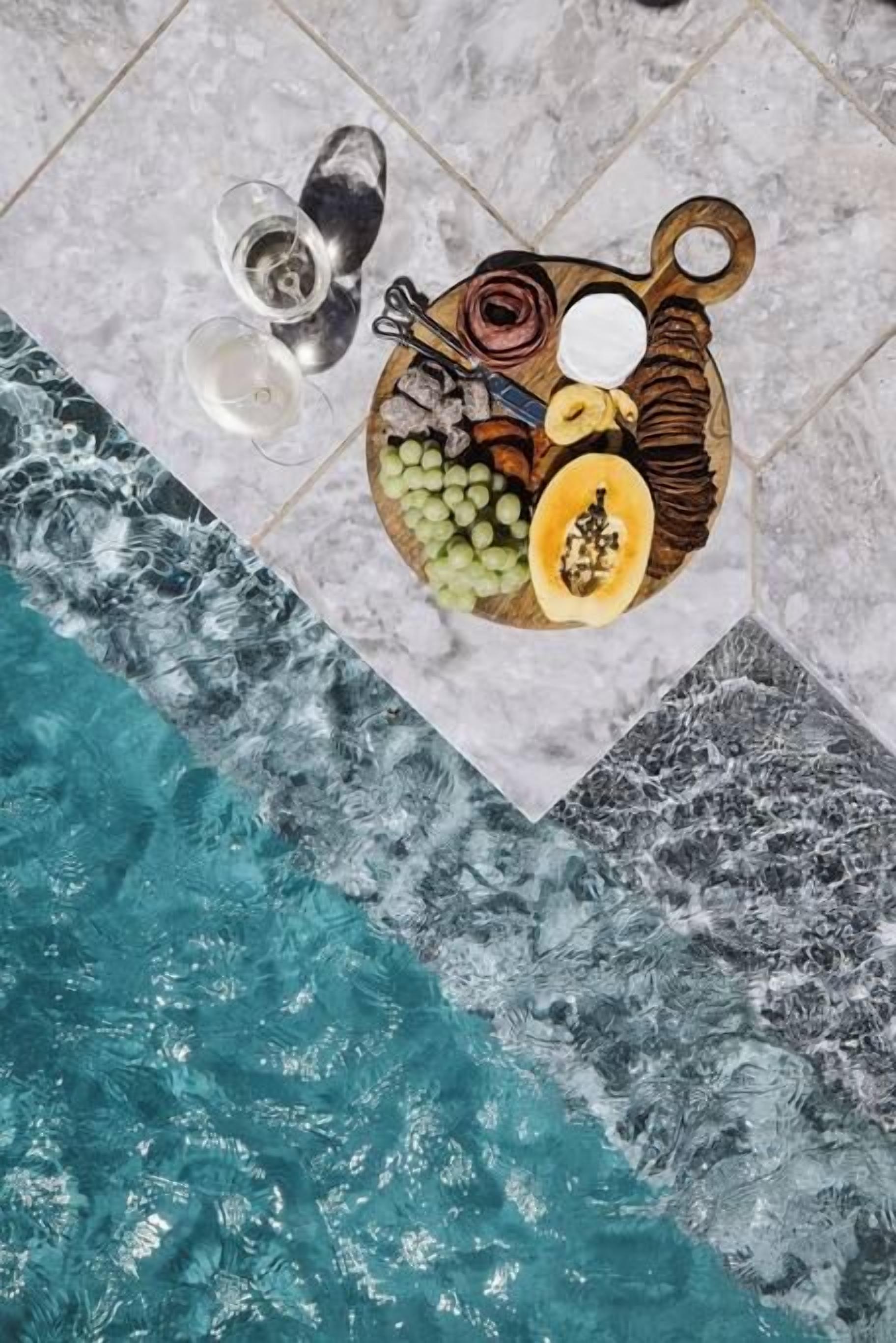 A poolside view with a fruit and cheese board, two glasses of water, and a towel.
