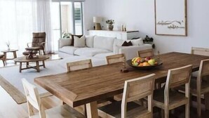 Un coin repas avec une table et des chaises en bois, un luminaire suspendu et un canapé blanc en arrière-plan.
