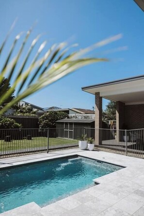 A swimming pool with a tiled deck, a covered patio area, and a fenced backyard.