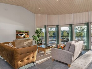 Un salon moderne avec un canapé, un fauteuil et une table basse, de grandes fenêtres avec des rideaux et un téléviseur fixé au mur.