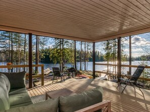 En overdækket terrasse med udendørs møbler, træloft og udsigt til søen.