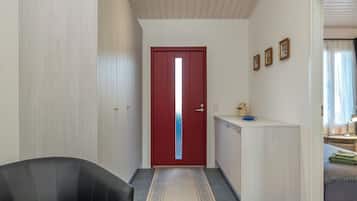 Un corridoio con una porta rossa, una sedia nera, un tappeto decorato, un armadietto bianco e un letto con un asciugamano verde.