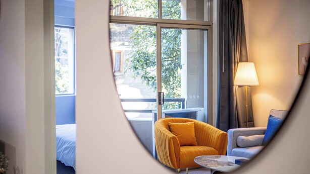 A hotel room with a bed, a yellow armchair, a grey sofa, a lamp, and a view of greenery outside.
