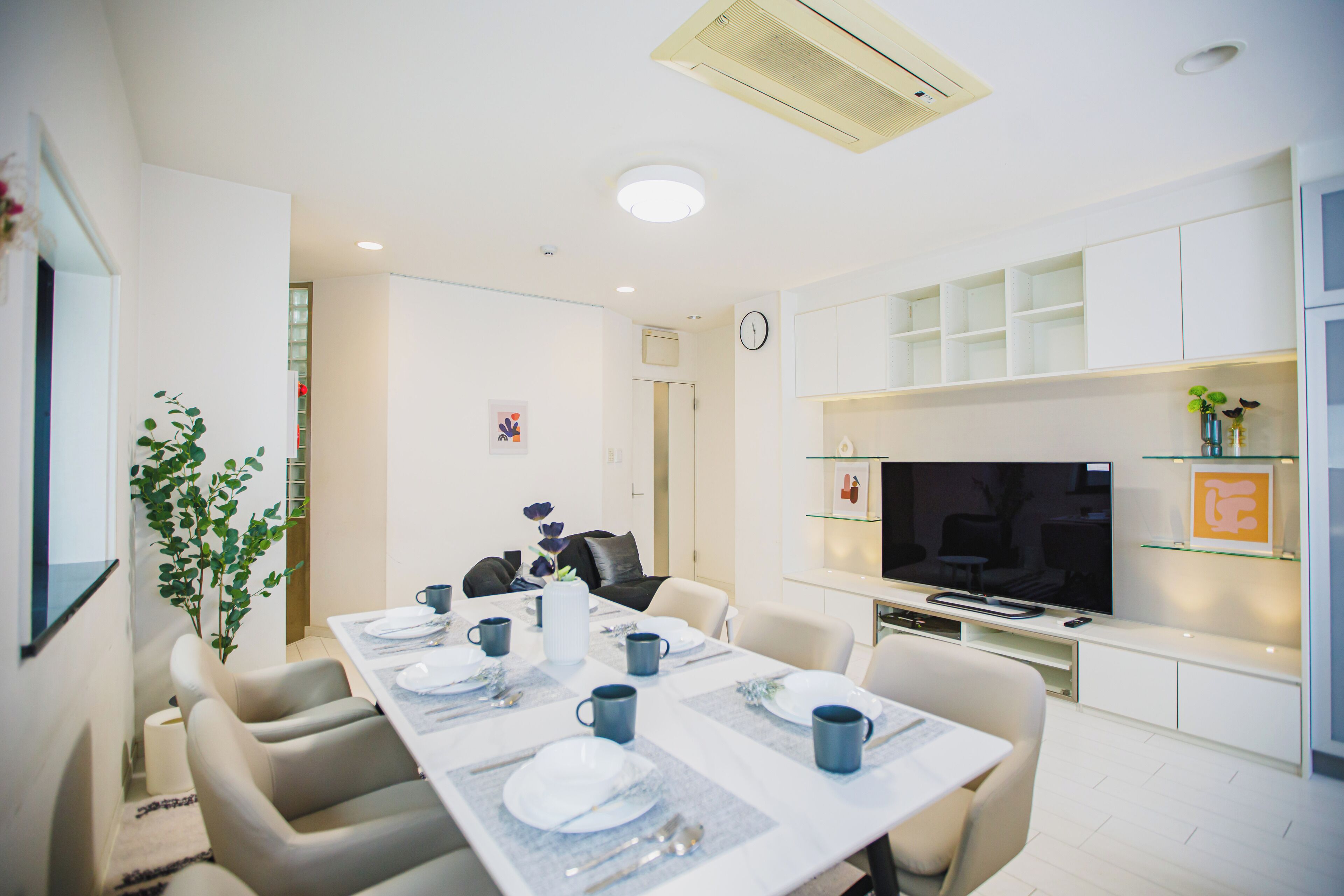 Un comedor moderno con una mesa larga dispuesta para cuatro personas, un televisor de pantalla plana y una planta pequeña.