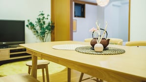 A wooden dining table with a placemat, a decorative vase with flowers, and a television in the background. - R Residence Tanimachi (Osaka)