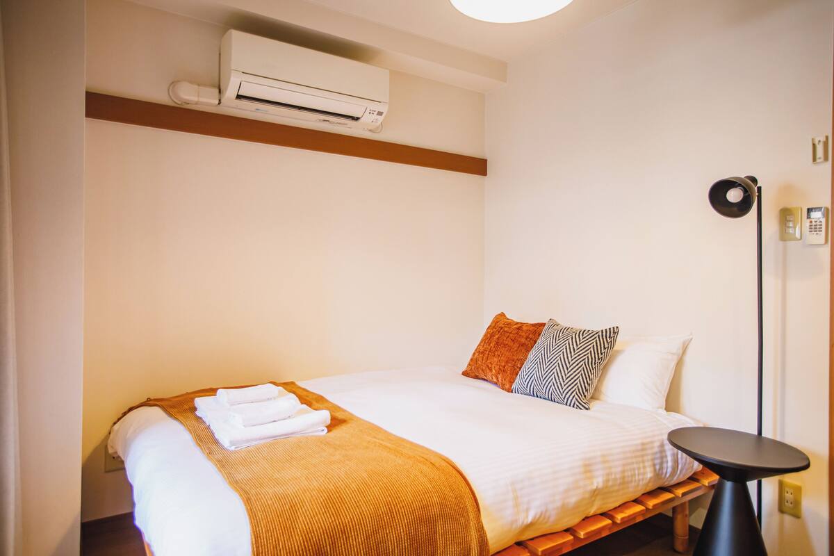 A hotel room with a bed, a bedside table, an air conditioning unit, a lamp, and a telephone.