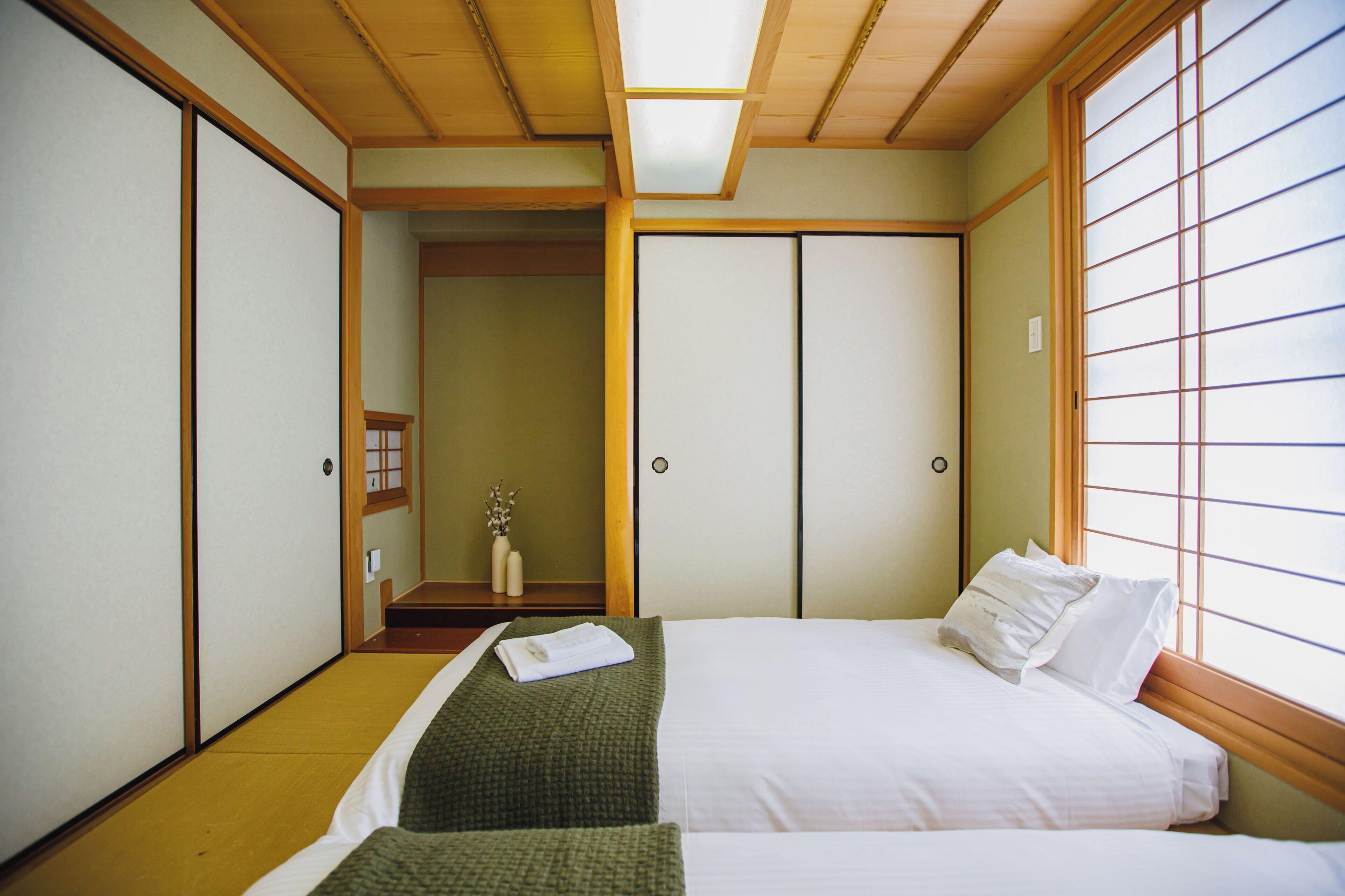 Perinteinen japanilaistyylinen makuuhuone, jossa on tatamimatto, futon-patja, liukuvat shoji-ovet ja näkymä shoji-seinän läpi.