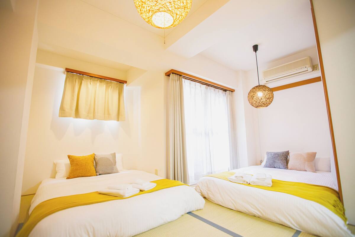 A hotel room with two beds, a ceiling light, an air conditioning unit, and a window with curtains.