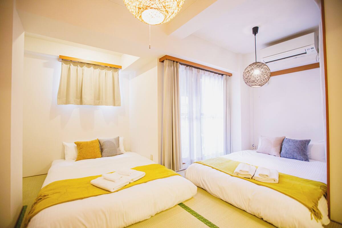 A hotel room with two beds, a window with curtains, a hanging light fixture, and an air conditioning unit.