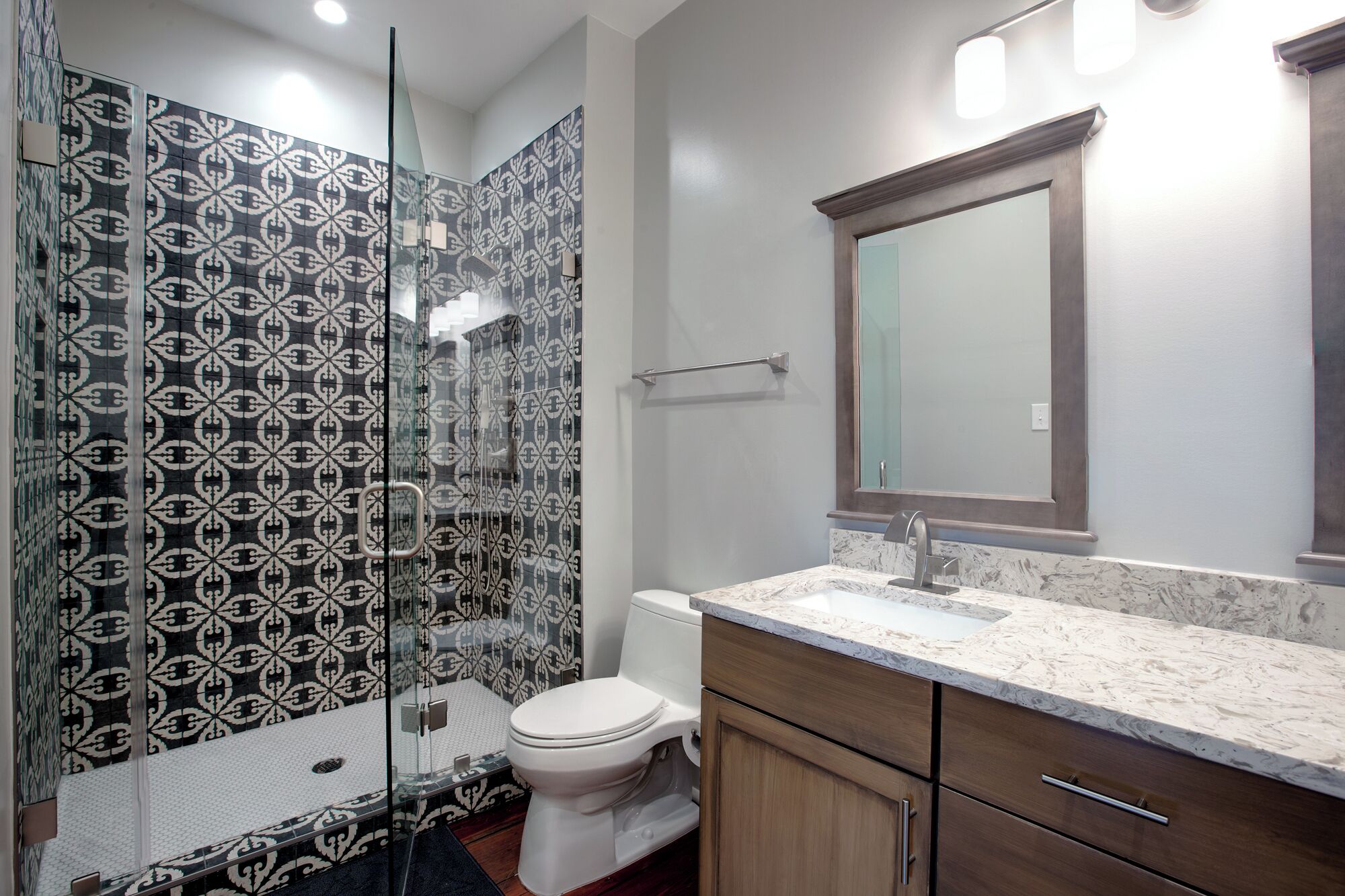 Un baño moderno con baldosas estampadas, cabina de ducha de vidrio, inodoro blanco, encimera de mármol y armarios de madera.