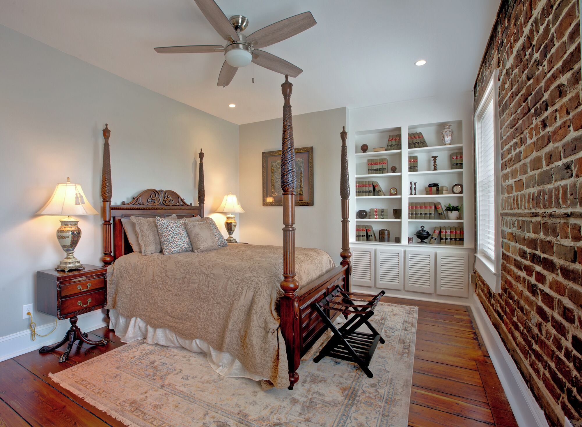 A four-poster bed with a ceiling fan, a bedside table with a lamp, and a built-in shelving unit with decorative items.