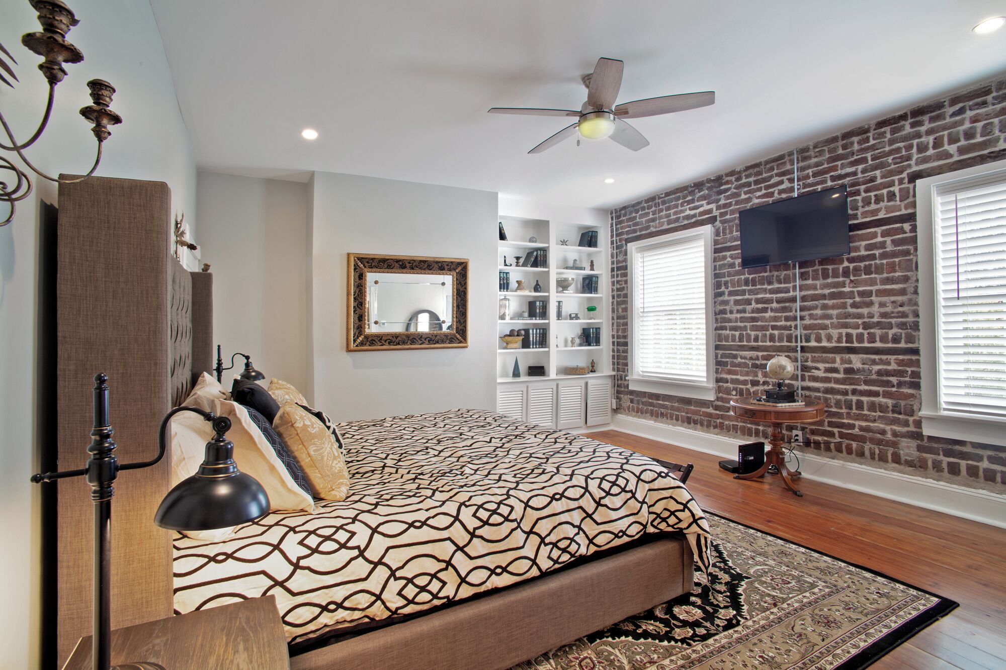 Une chambre à coucher avec un lit, un ventilateur de plafond, une bibliothèque et un téléviseur fixé à un mur en briques.