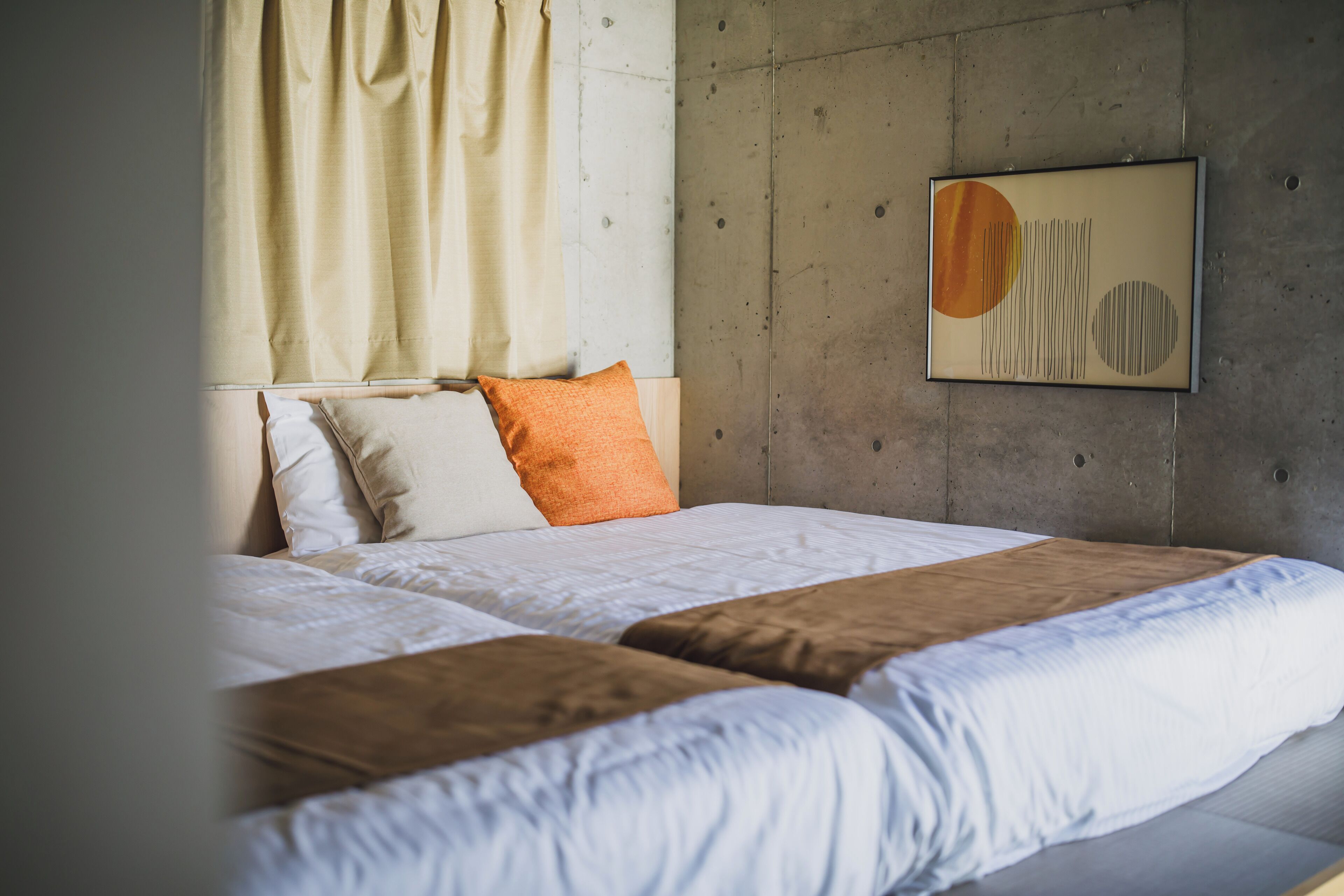 Cama com roupa de cama branca e marrom, travesseiro bege e uma obra de arte abstrata na parede.