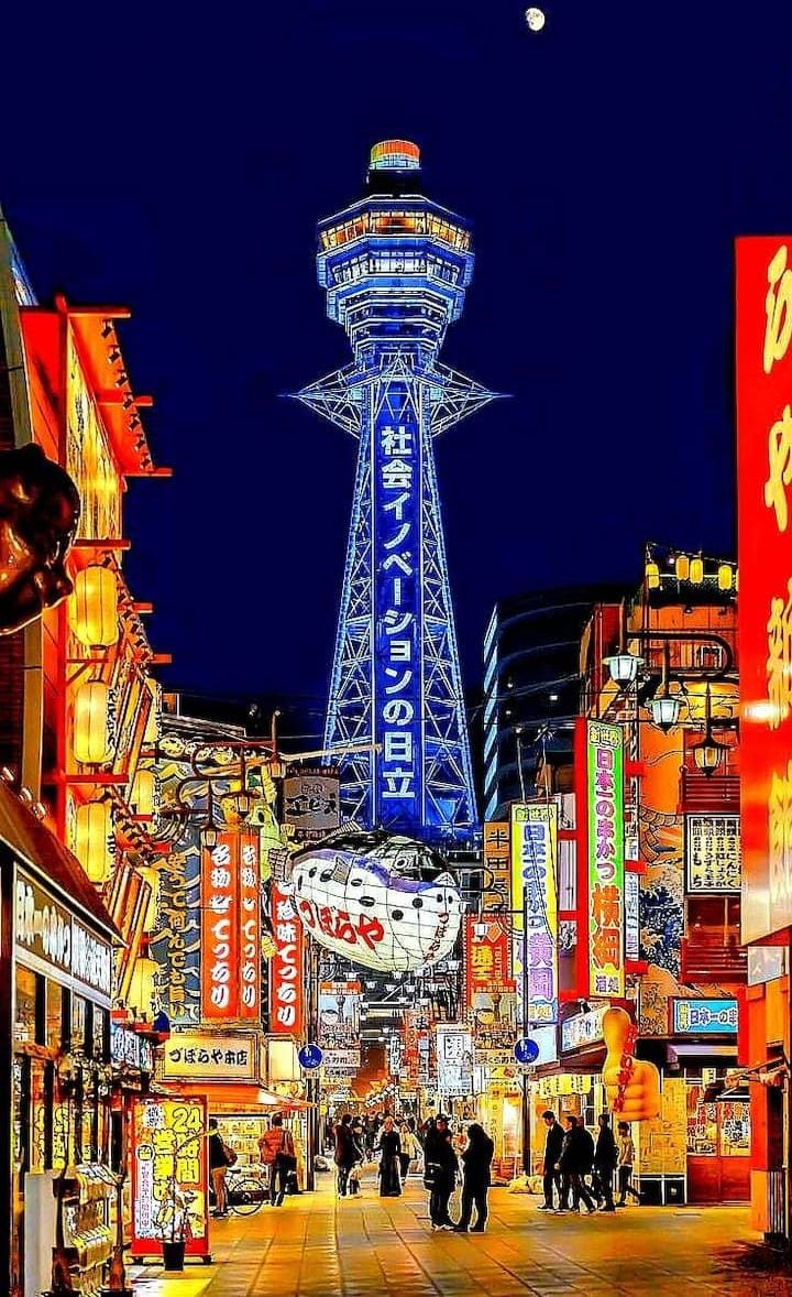 A busy urban street at night with illuminated signage and a prominent tower structure.