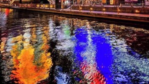 Une zone urbaine aux éclairages vifs, avec des panneaux publicitaires illuminés et des reflets sur l’eau.