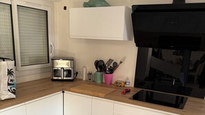 A modern kitchen with white cabinets, a stainless steel range, a microwave, and a coffee maker. There is a window with blinds, a pink mug with utensils, and a small trash bin. - House 10 min from the sea With garden and swimming pool (Fréjus)