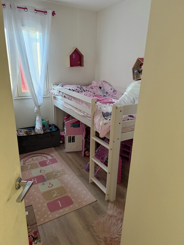 Une chambre d’enfant avec des lits superposés, une petite maison de jeu et une fenêtre ornée de rideaux légers.