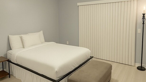 A bedroom with a bed, an ottoman, a nightstand, a floor lamp, and a window with blinds. - Cozy 2BR 2BA on Fremont street 3 min walk from Fremont experience. (Las Vegas)