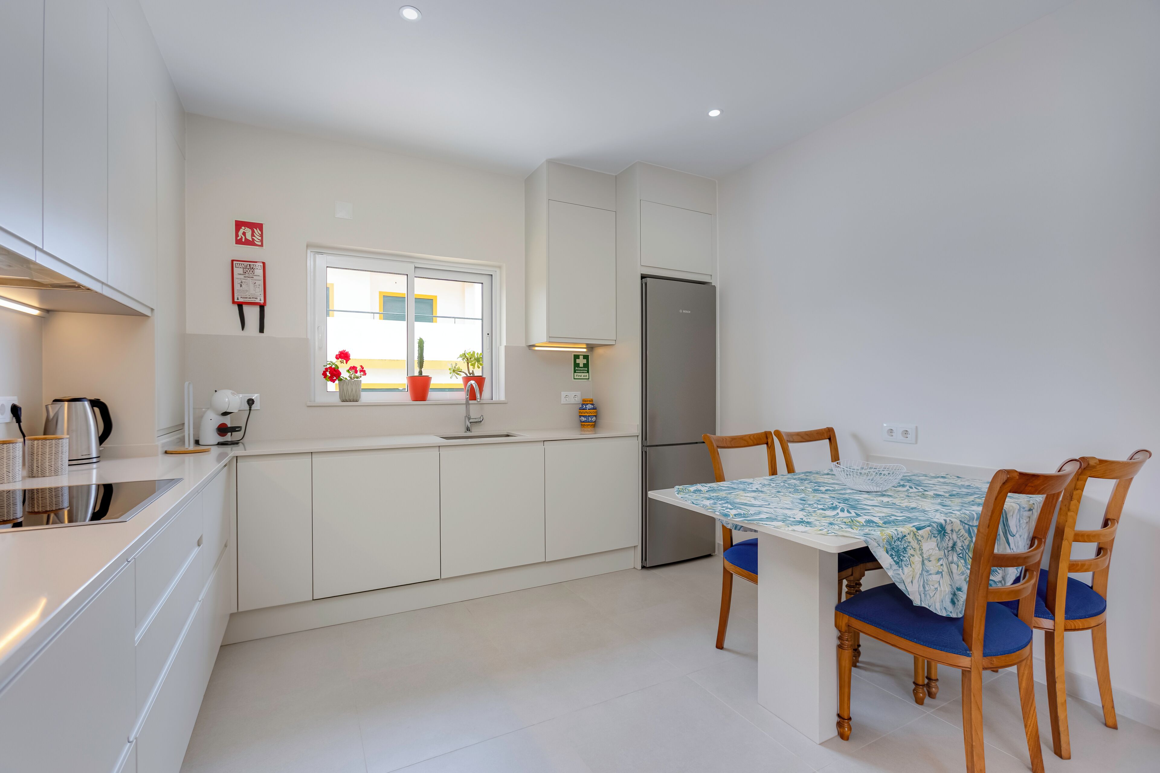 A modern kitchen with white cabinets, a stainless steel refrigerator, a dining table with blue chairs, and a window with potted plants.