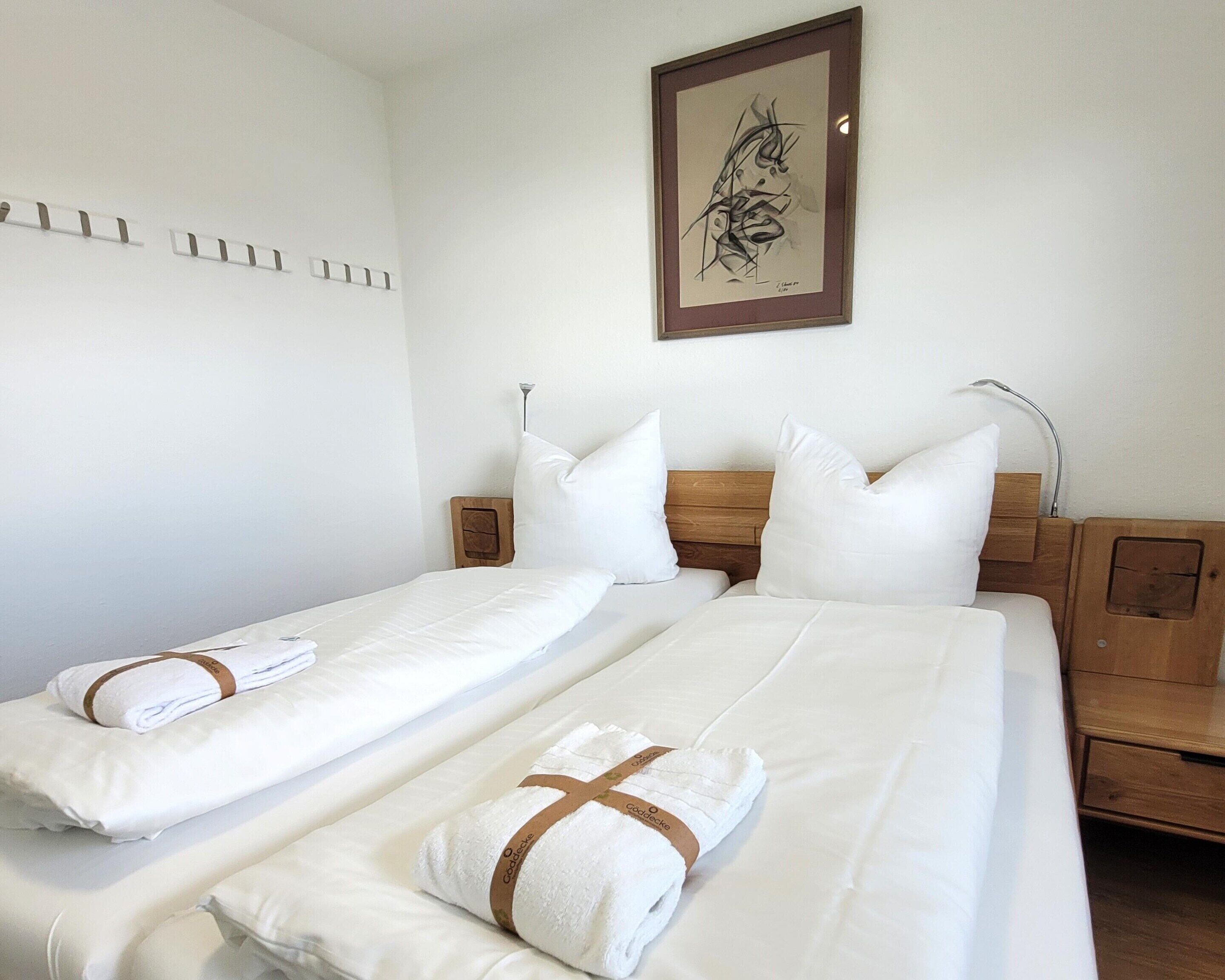 Ein Hotelzimmer mit zwei Betten, jeweils mit weißen Bettwäsche und Handtüchern, einem Holz-Kopfteil, einem Nachttisch und einem gerahmten Kunstwerk an der Wand.