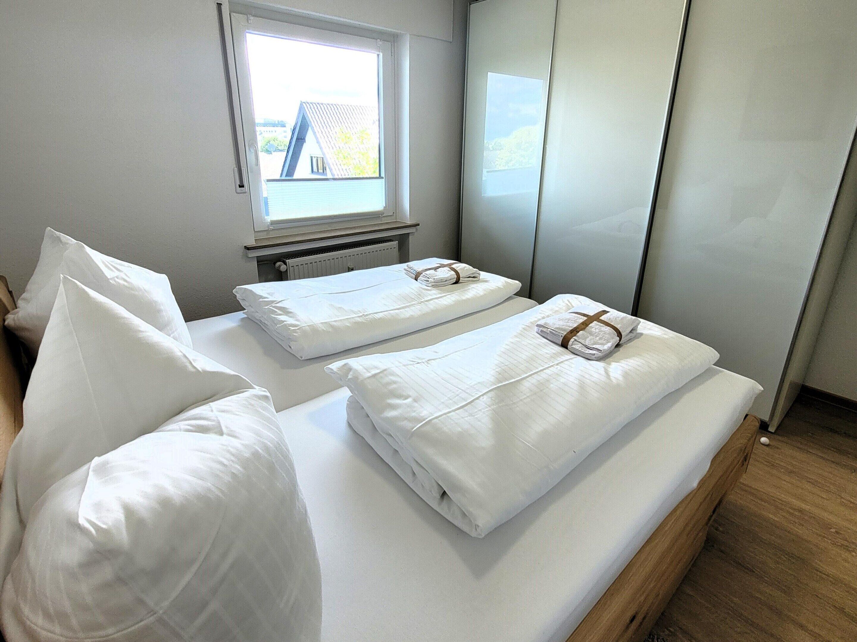 Habitación de hotel con dos camas, cada una con ropa de cama y toallas blancas, una ventana con vista a edificios y un radiador debajo de la ventana.