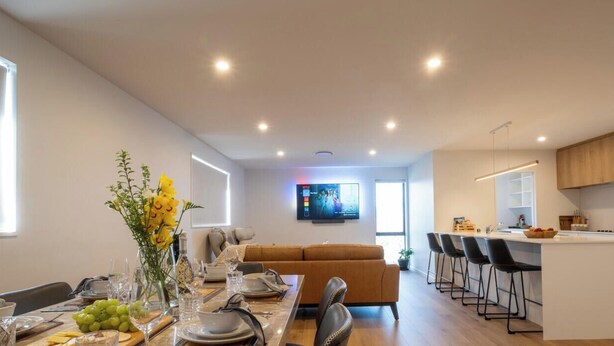 Eine moderne Küche mit einer zentralen Kochinsel, ein Essbereich mit einem langen Tisch für sechs Personen und ein Wohnbereich mit Sofa und Fernseher.